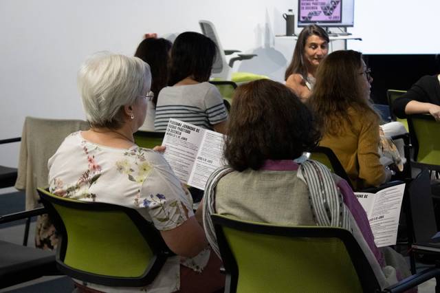 A group of people sitting in a seminar room, listening to a speaker while holding printed handouts.