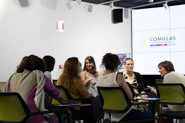 A group of women engaging in a discussion in a classroom with a projector displaying the 'Comillas Pontifical University' logo.
