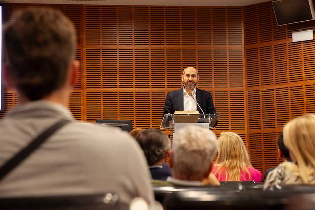 A man speaks at a podium in front of an audience in a room with wooden wall paneling.