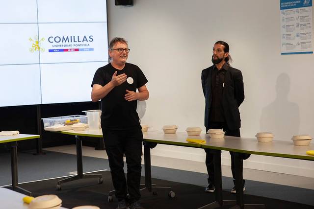 Two men presenting in a room with a table displaying multiple white circular objects, under a sign reading 'COMILLAS UNIVERSIDAD PONTIFICA'.