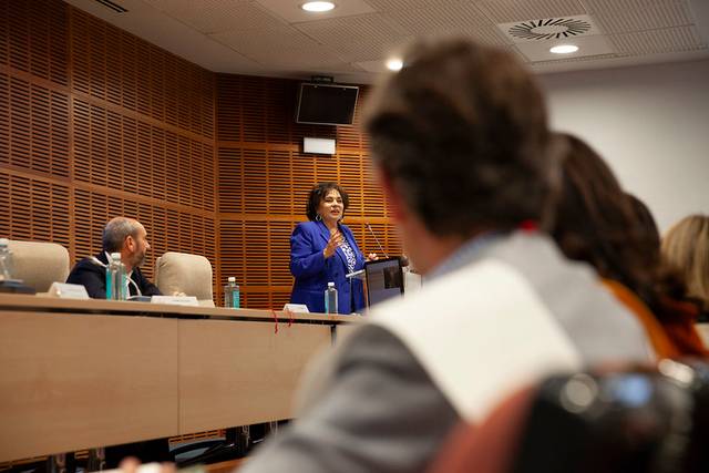 A woman in a blue suit speaks at a podium in a conference room with listeners seated in front.