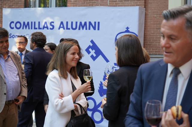 A group of people socializing at an event with a 'Comillas Alumni' banner in the background.