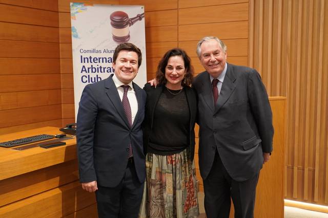 Three people smiling for a photo inside a wooden paneled room with a sign titled 'Comillas Alumni International Arbitration Club'.