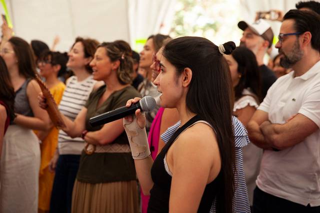 A young woman sings at a microphone in front of an audience under a tent.