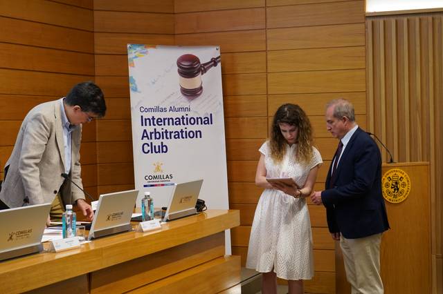A man and a woman are looking at a tablet in a conference room with a banner reading 'Comillas Alumni International Arbitration Club'.