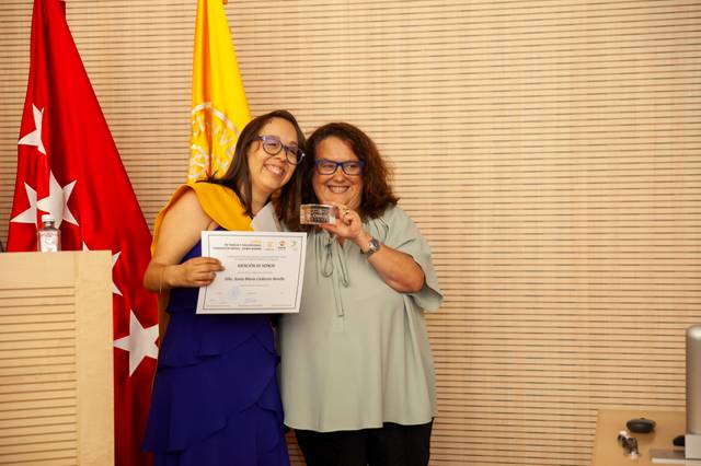 Two women smiling and holding a certificate together in a room with flags in the background.