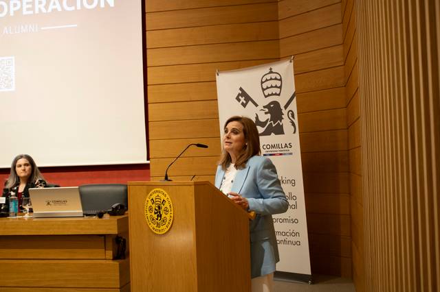 A woman is giving a speech at a podium in a conference room with banners and a projector screen in the background.