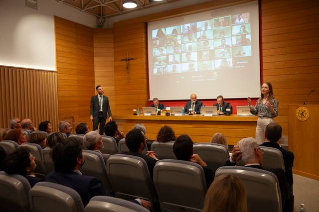 A woman presenting in a conference room with an audience and virtual participants displayed on large screens.