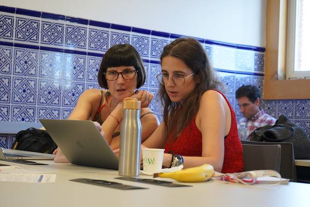Two women discussing over a laptop in a room with blue tile walls.