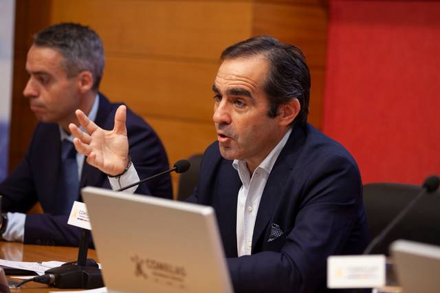 A man in a dark suit is speaking and gesturing during a meeting, with another man partly visible in the foreground.