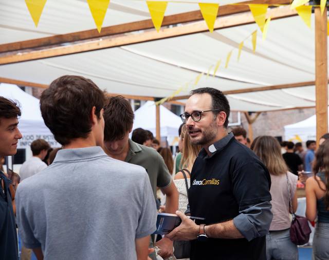 A man wearing glasses and a black t-shirt with a logo is engaging in a conversation with three young people at an outdoor event under a canopy decorated with yellow triangular flags.