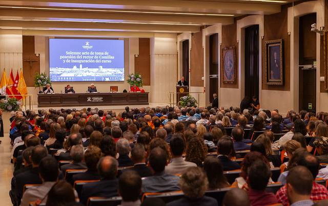 A formal event in a large hall with audience members attentively listening to a speaker at the podium.