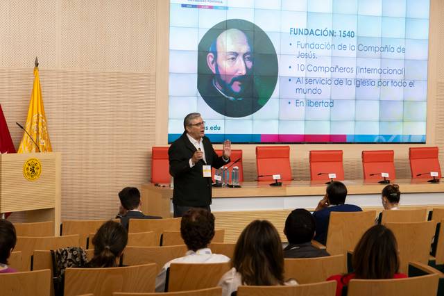 A man presents a lecture to an audience in a modern conference room with a presentation slide featuring historical content in the background.