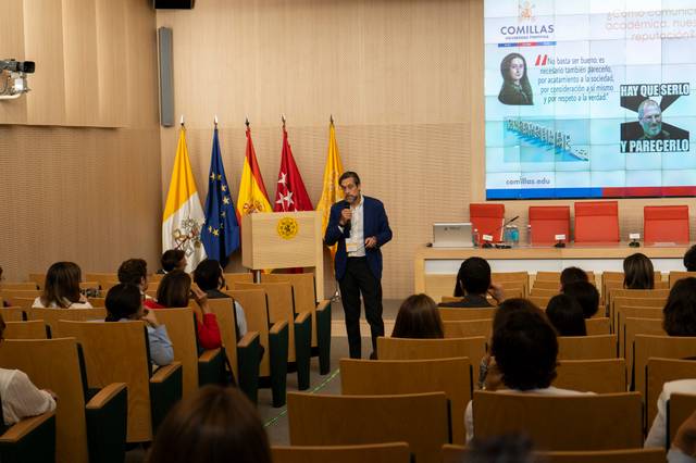 A man is presenting a lecture in a university auditorium with students and flags in the background.