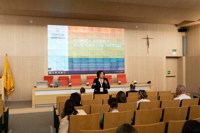 A woman is presenting at a university lecture hall in front of an audience, with a large screen behind her displaying text about alumni and corporate relations.