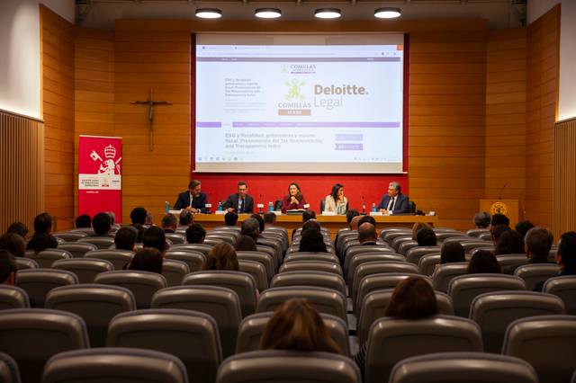 Panel of speakers at a corporate event in a conference hall with audience members seated, facing a screen displaying Deloitte and Comillas Legal logos.