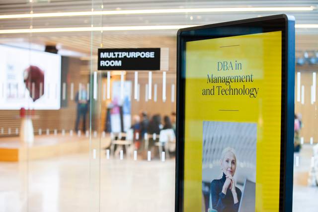 An indoor informational display showing an advertisement for a DBA in Management and Technology program, with a blurred view of a lobby in the background.