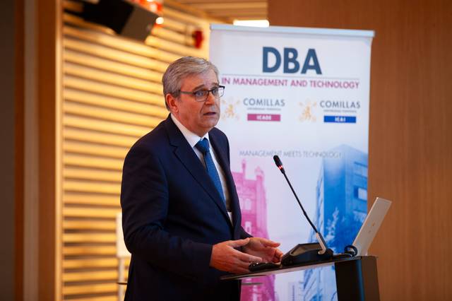 A man in a suit speaking at a podium with a 'DBA in Management and Technology, Comillas' banner in the background.