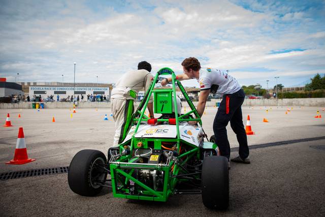 Two people work on a small green race car in a parking lot marked with orange cones.