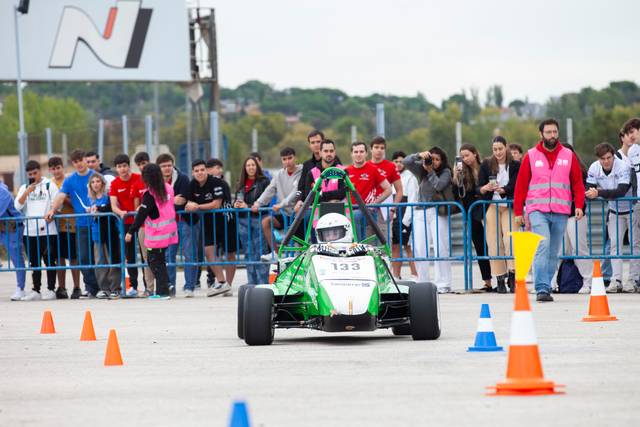 A small green racing car competes in a cone-lined track with spectators watching from behind barriers.