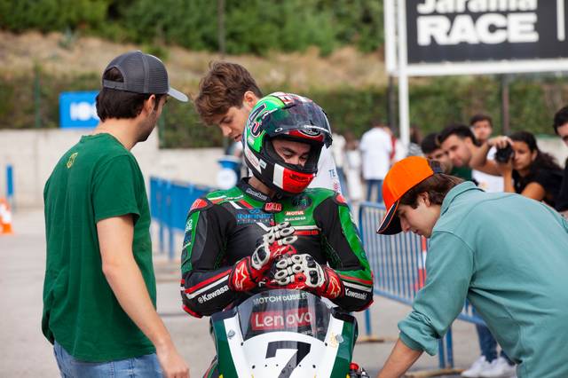 A motorcyclist in a full racing suit and helmet is surrounded by three young men at an outdoor racing event.