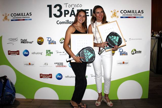 Two women holding paddle tennis rackets pose with their trophies at a promotional event.