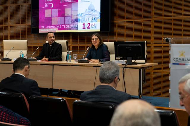 A man and a woman are seated at a conference table in front of an audience, with a presentation about theology on a screen behind them.