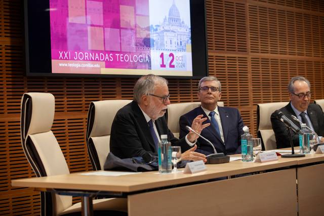 Three men sitting at a panel discussion during an event with a background banner displaying 'XXI Jornadas Teologia'.