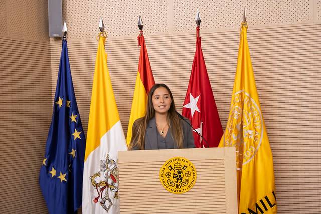 A woman is giving a speech at a podium with multiple flags behind her, including the European Union and various regional flags.