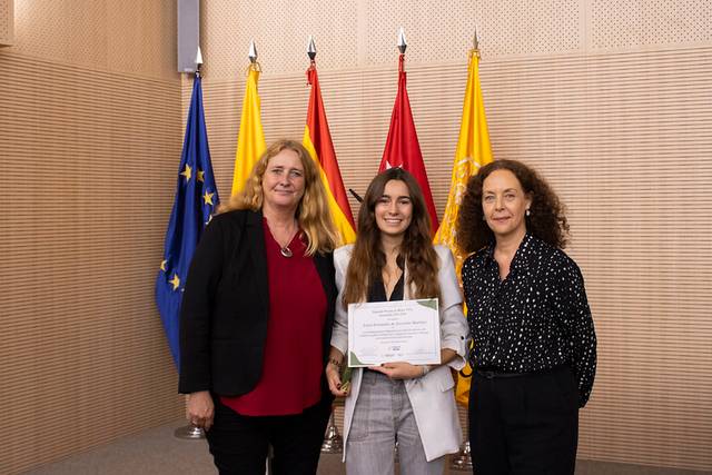 Three women pose together during an award ceremony where the middle woman holds a certificate, with flags in the background.