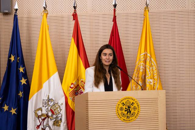 A woman is giving a speech at a podium with multiple flags, including the European Union and Spanish flags, in the background.