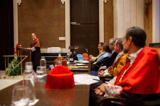 A speaker presenting at a formal academic event with attentive audience members dressed in ceremonial robes.