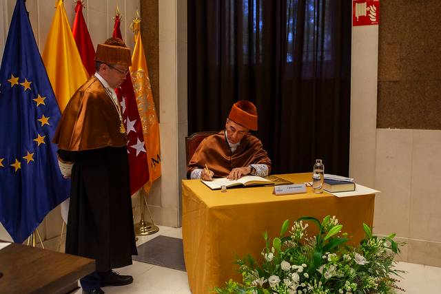 A man in traditional Moroccan attire is signing a document at a table, while another man in a formal suit watches.