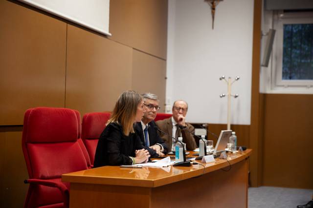 Three people are seated at a table at a business or academic seminar, facing an audience, with red chairs and microphones visible.