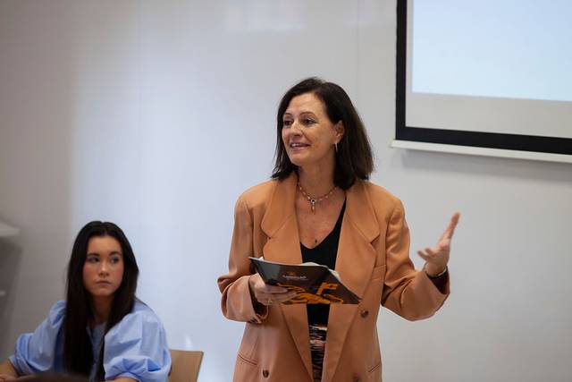 A woman stands teaching in front of a classroom, gesturing with one hand while holding a tablet, with a young woman looking on from the background.