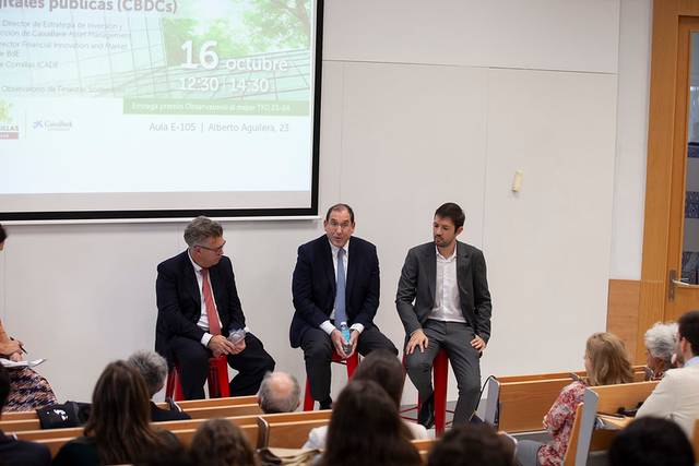 Three men sit on a stage in a conference hall during an event about central bank digital currencies.