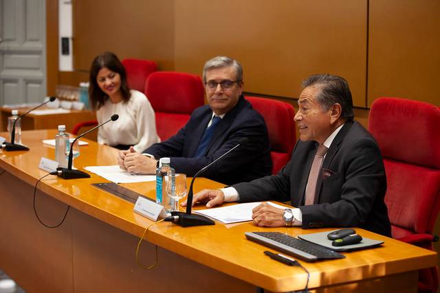 Three people sitting at a conference table in a formal setting, the middle man is speaking.