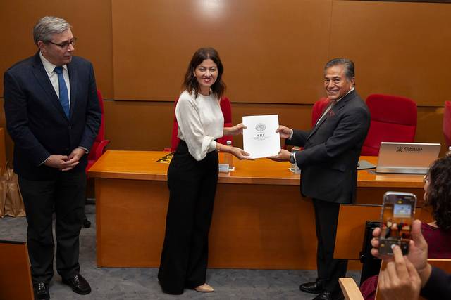 A woman is standing between two men, receiving a document in a formal indoor ceremony while another person takes a photo.