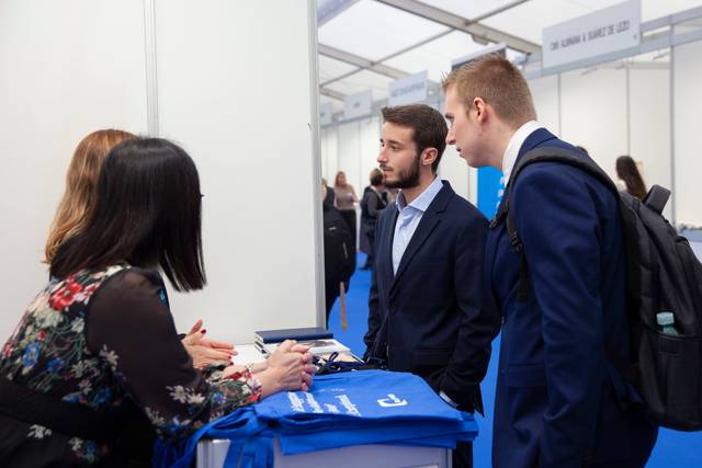 Two men in business attire talking to a woman at a registration desk during an event.