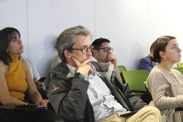 A group of adults attentively listening to a speaker at a seminar or lecture, sitting on green chairs in a modern room.