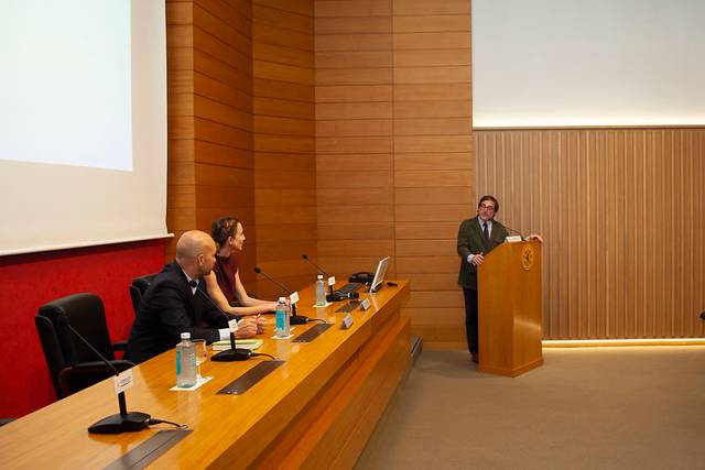 Un hombre está de pie hablando en un podio ante tres personas sentadas en una mesa durante una conferencia en una sala con paredes de madera.