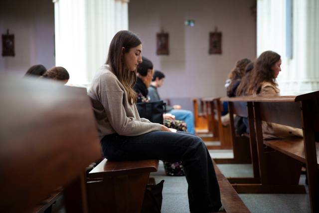 A young woman sitting thoughtfully in a church pew while other people are seated around her.