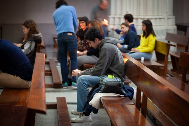 A young man is sitting alone on a wooden bench, looking at his phone, while other people sit and interact in the background inside a spacious hall.