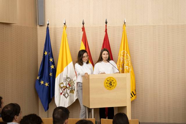 Two women speaking at a podium with various national flags in the background, including the flags of the European Union and Spain.