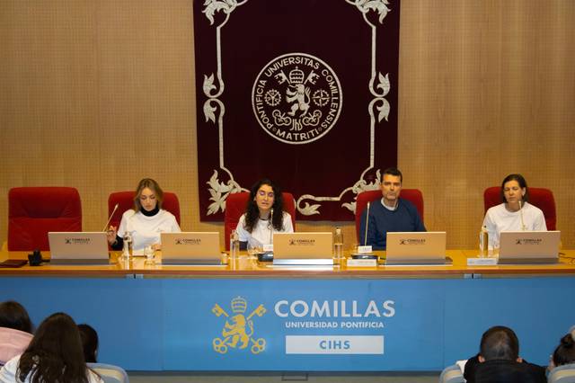 Four people are seated at a conference table in a university setting, each with name tags and microphones, in front of an attentive audience.