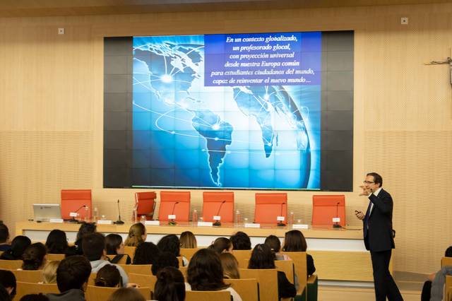 A man presenting in a conference room with a digital screen displaying a global map and text behind him.