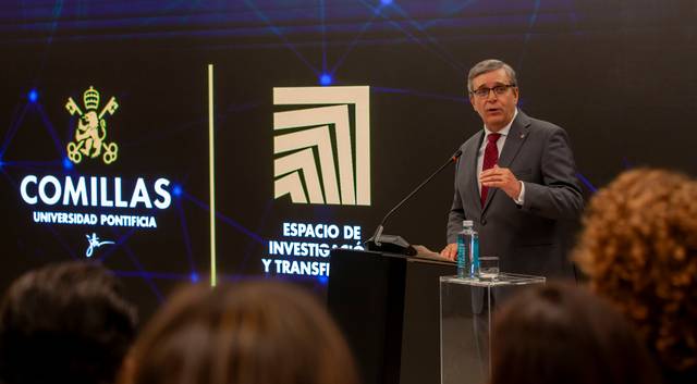 A man giving a speech at a lectern with 'Comillas Universidad Pontificia' and 'Espacio de Investigación y Transfi' logos in the background.