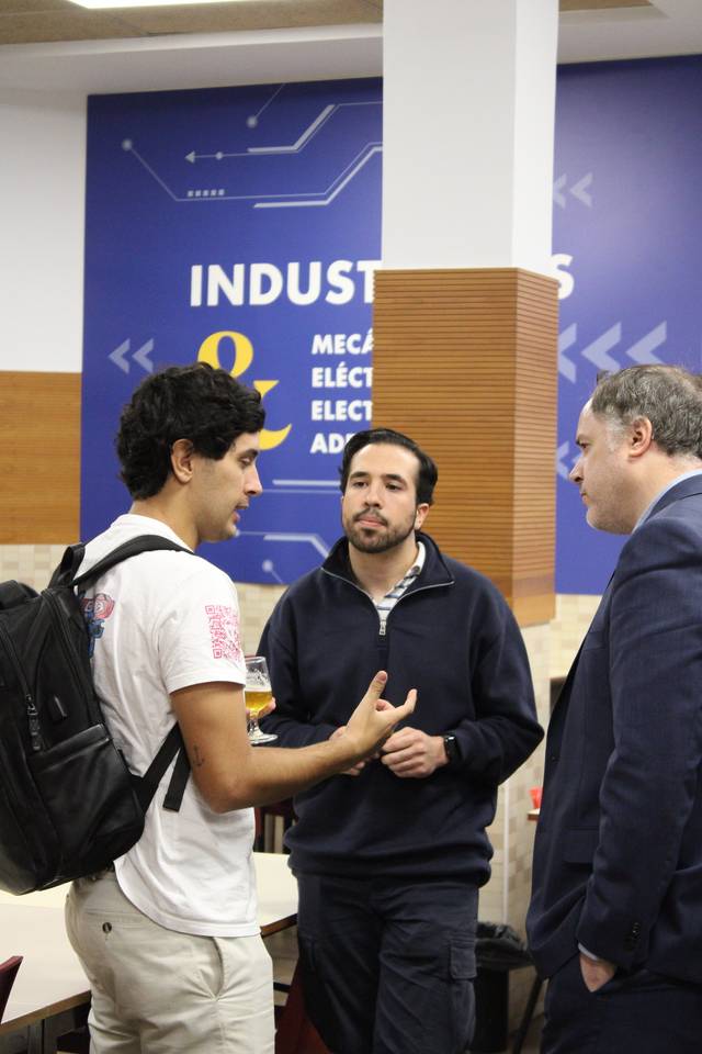 Tres hombres conversan en un pasillo con un cartel de 'Industria Mecánica, Electrónica y Eléctrica' al fondo.