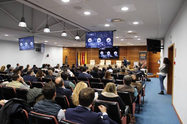 Una conferencia en un salón con numerosos asistentes sentados, escuchando a un hablante en un podio.