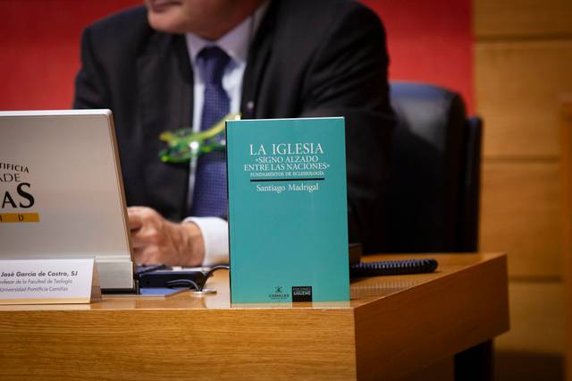 Un hombre sentado tras un escritorio con un libro titulado 'La Iglesia Signo y Azote Entre las Naciones' visible delante de él.